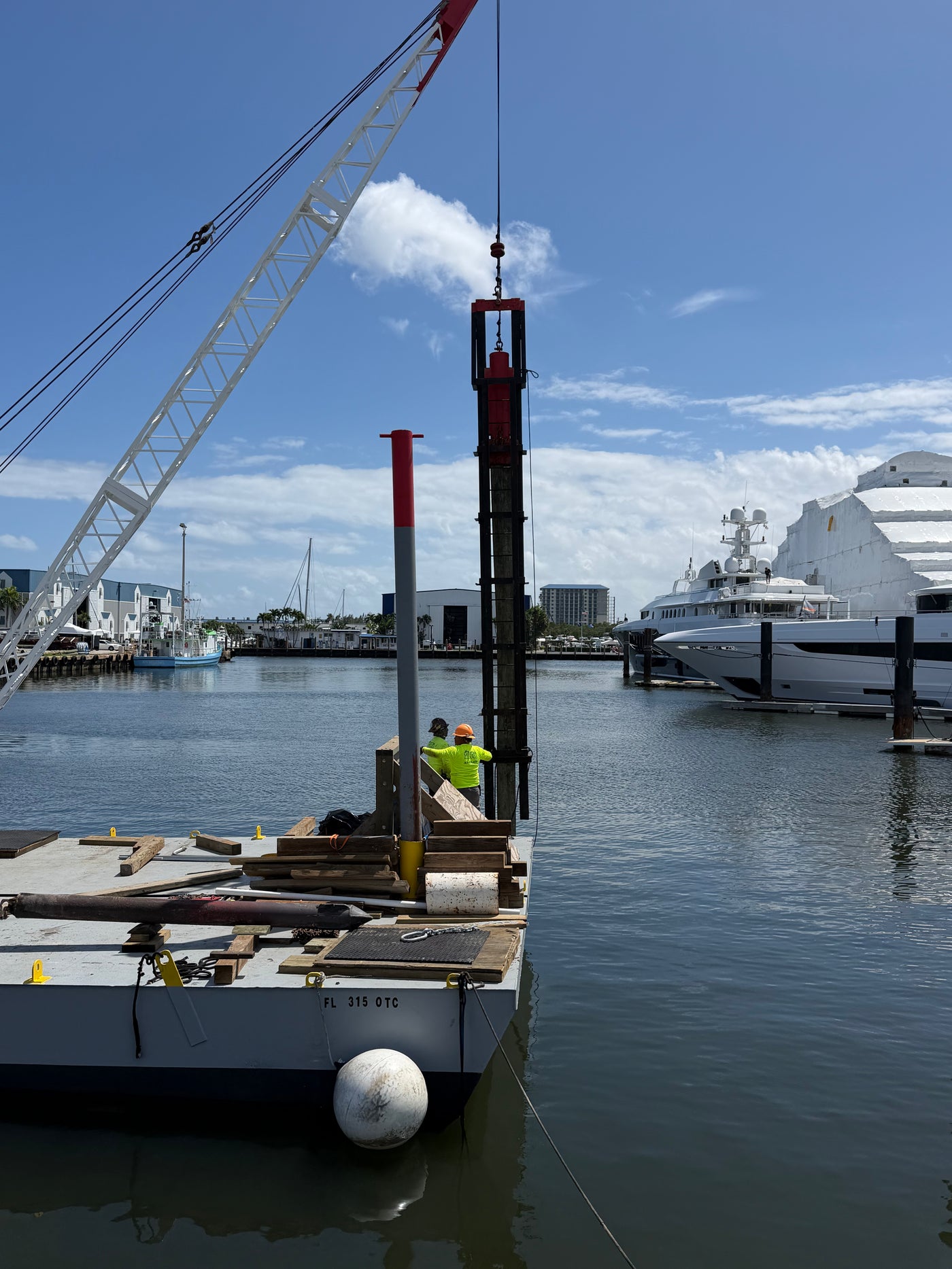 Pile-driving rig installing a marine foundation pile from a work barge, with a yacht and marina in the background.